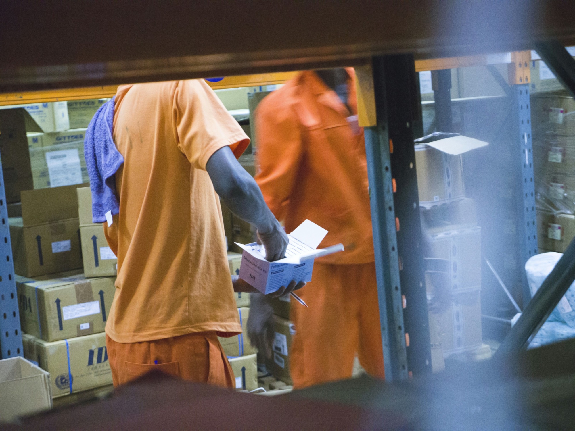 Two warehouse workers handling boxes of medical supplies between racks, a shelf blurred in front