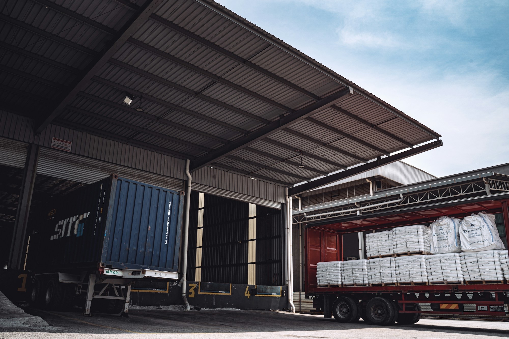 A container trailer and a curtain-side truck loaded with bagged material at a warehouse loading dock