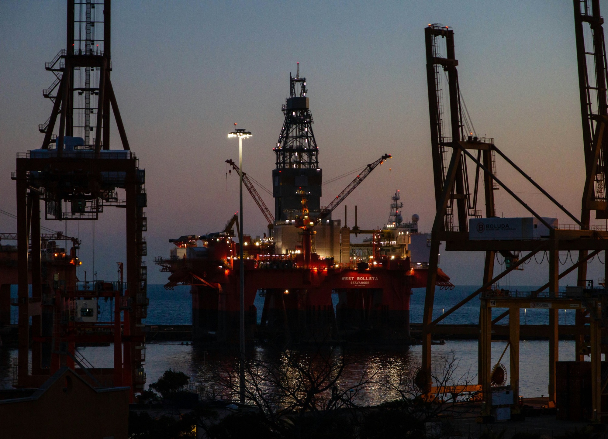 A drilling rig lit up in harbour at dusk, framed by quayside cranes