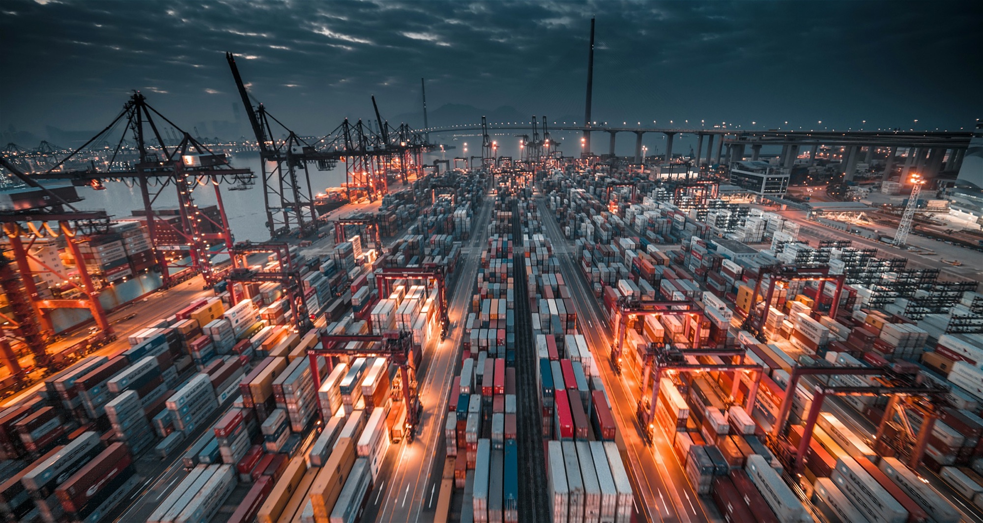 A container terminal at dusk, lit yard cranes over stacked rows and light trails from trucks